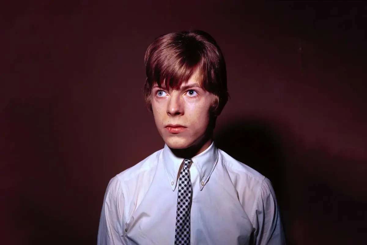 David Bowie posing in an early-career studio portrait, wearing a white button-down shirt and patterned tie, looking off to the side with a serious expression against a dark background.
