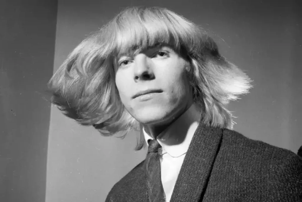 David Bowie captured in a black-and-white portrait with longer, windswept hair, wearing a suit jacket and tie, gazing calmly at the camera in a mid-1960s era photo.