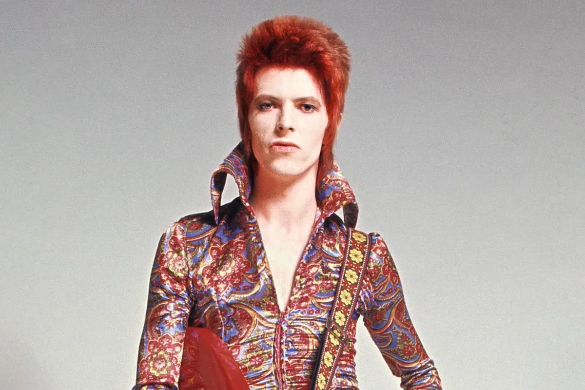 David Bowie posing in a classic Ziggy Stardust-era studio portrait, wearing a colorful patterned jumpsuit with a high collar and bright red hair, standing confidently against a neutral background.