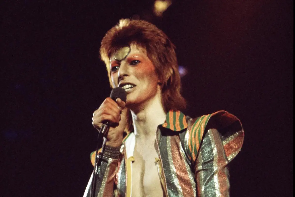 David Bowie in a colorful, patterned stage outfit sings into a microphone under dramatic stage lighting.