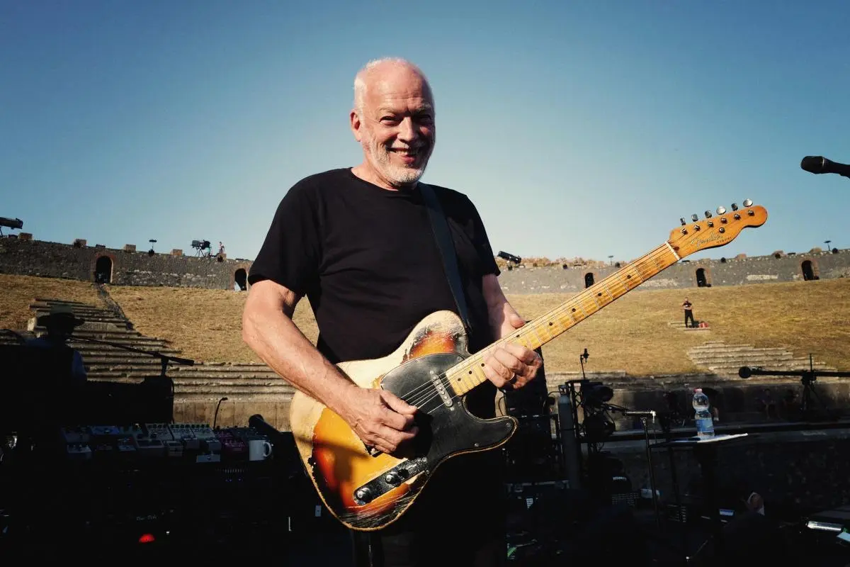 David Gilmour A smiling guitarist holds a worn electric guitar while standing on an outdoor stage set against ancient stone seating under a clear sky.