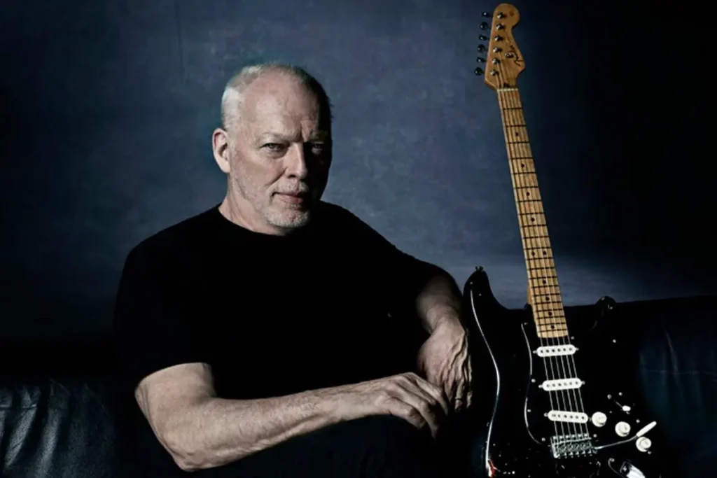 David Gilmour sits in a dark studio beside a black electric guitar, looking calmly toward the camera with folded hands.