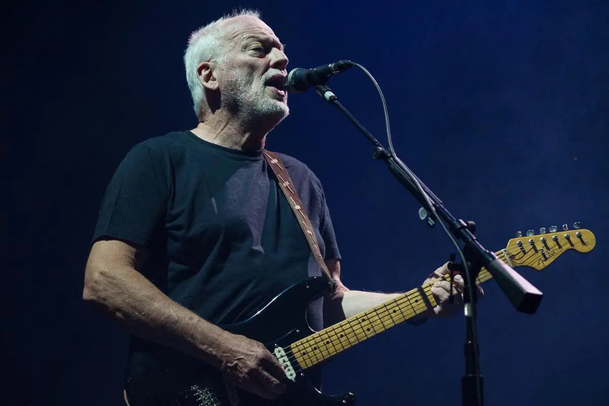 David Gilmour sings into a microphone while playing an electric guitar under dim blue stage lighting.