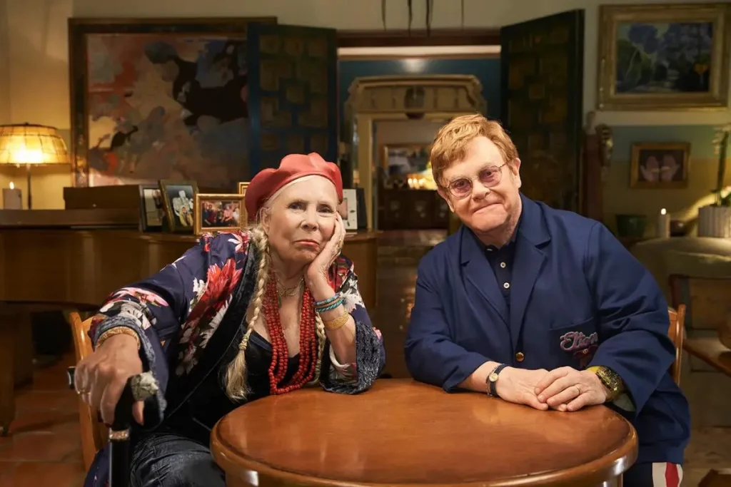 Joni Mitchell and Elton John, sit together at a wooden table in a warmly lit room.
