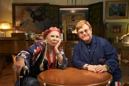 Elton John & Joni Mitchell: The Quiet Alliance That Bent Rock Sideways Joni Mitchell and Elton John, sit together at a wooden table in a warmly lit room.