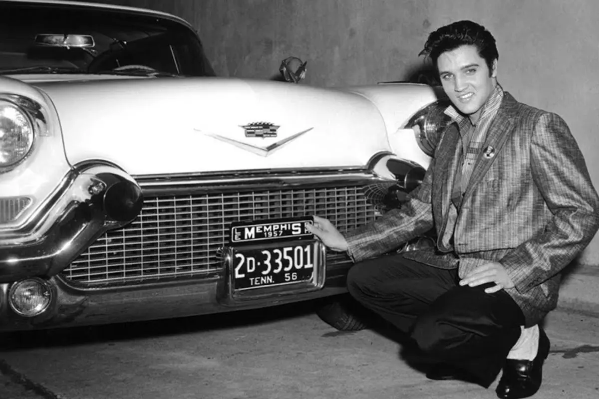 Elvis Presley in a plaid jacket kneels beside a classic car, smiling as he holds up its license plate in a garage or indoor setting.