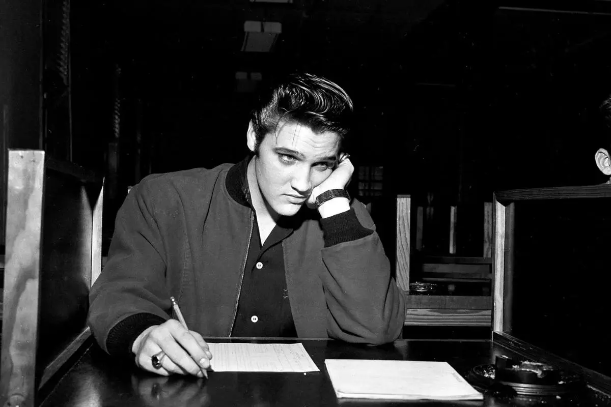 Elvis Presley with styled hair sits at a desk, leaning his head on one hand while writing on a form, appearing thoughtful or tired in the dimly lit room.