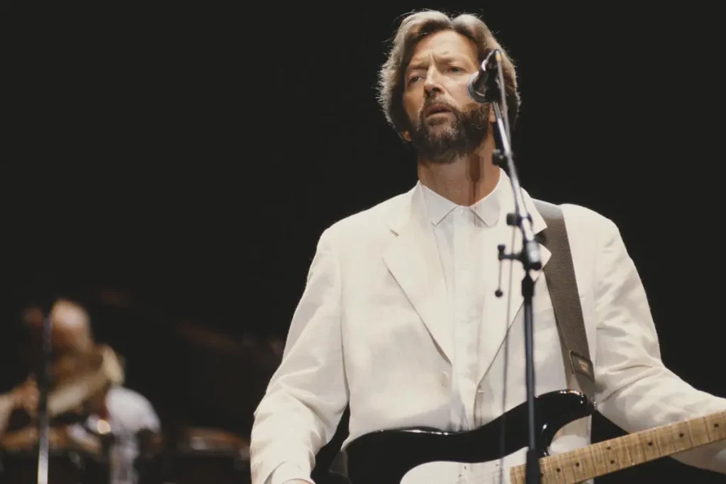 Eric Clapton performs onstage wearing a white suit and playing a black-and-white electric guitar.