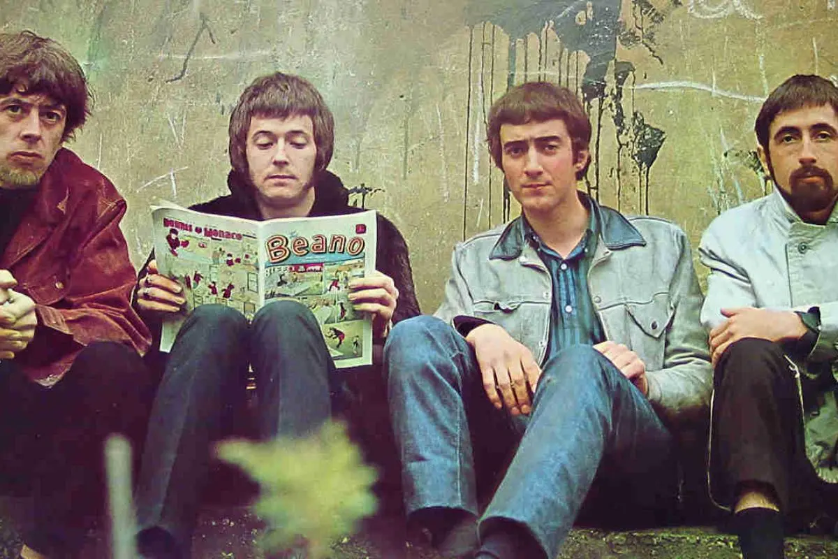 Bluesbreakers with Beano sitting against a worn, graffiti-marked wall, with one reading a Beano comic while the others look toward the camera.