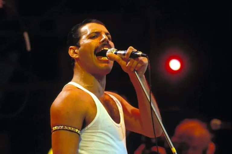 Freddie Mercury performing onstage wearing a white tank top and gold armband.