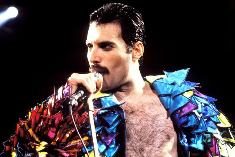 Freddie Mercury performs onstage in a colorful, sculpted jacket, holding a microphone and singing passionately.