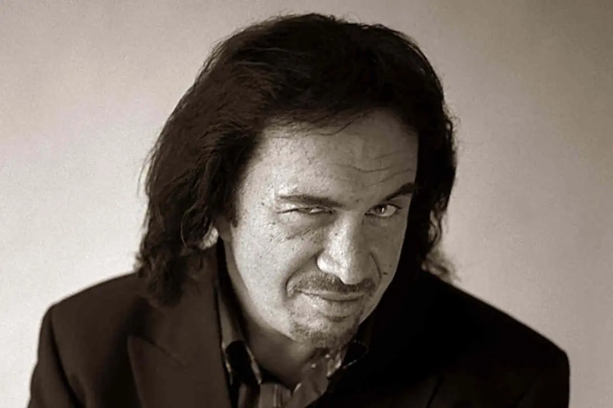 Gene Simmons in a suit jacket, giving a playful wink while slightly smirking.