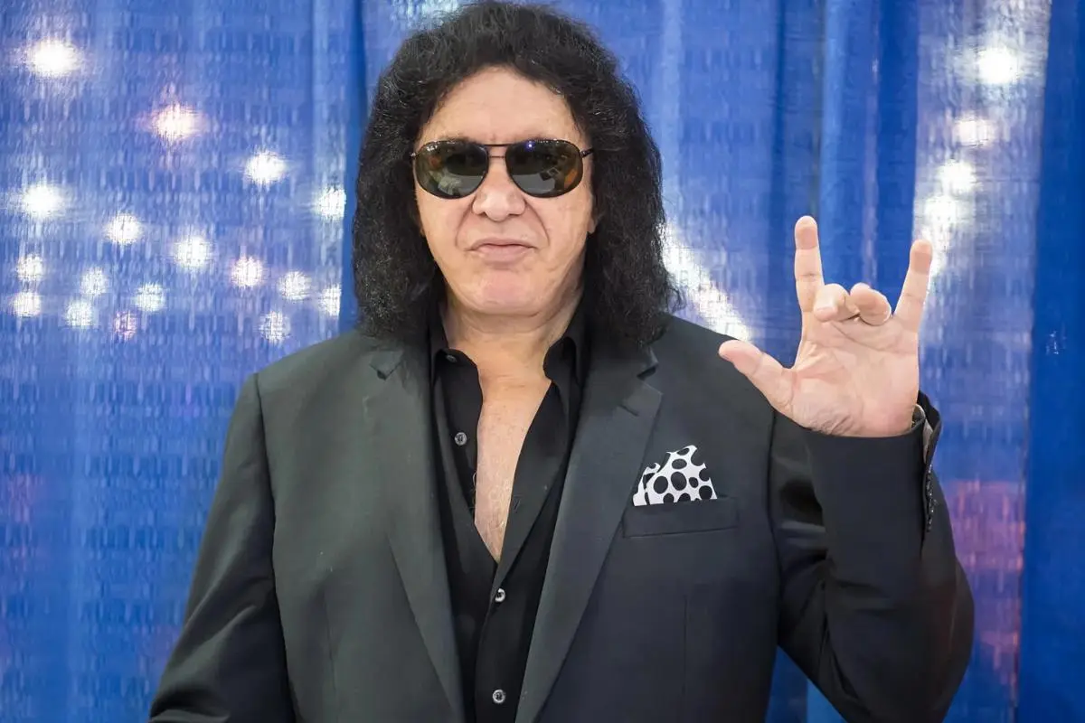 Gene Simmons wearing a black suit and dark sunglasses, standing in front of a blue backdrop.