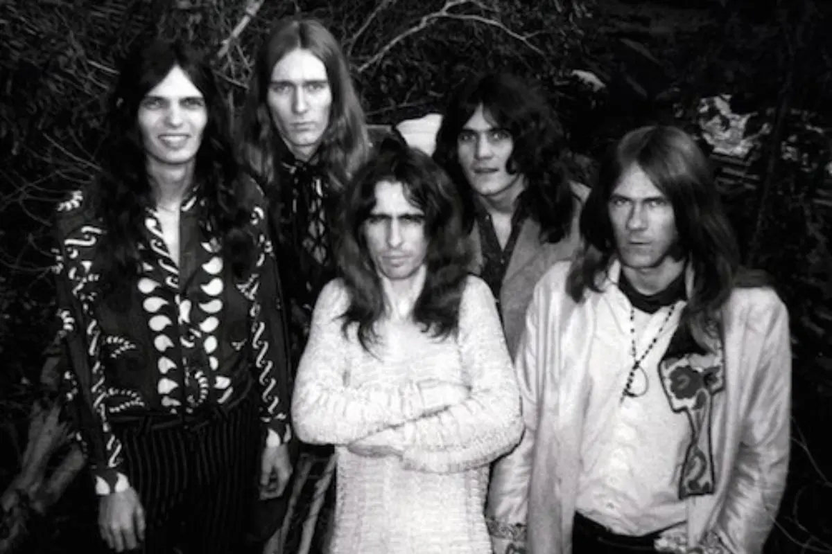 Alice Cooper band stand outdoors, wearing patterned and vintage-style clothing, with the man in front crossing his arms and staring intensely at the camera.