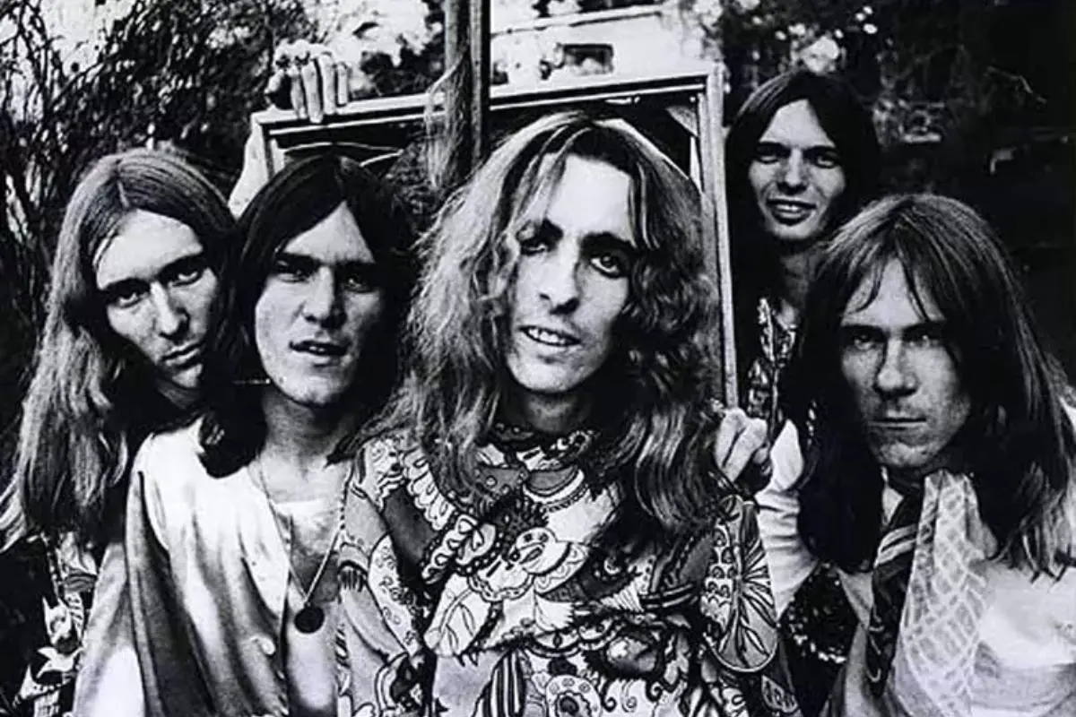 Alice Cooper band with long hair pose closely together outside, wearing eclectic, patterned outfits as they look directly at the camera with serious expressions.