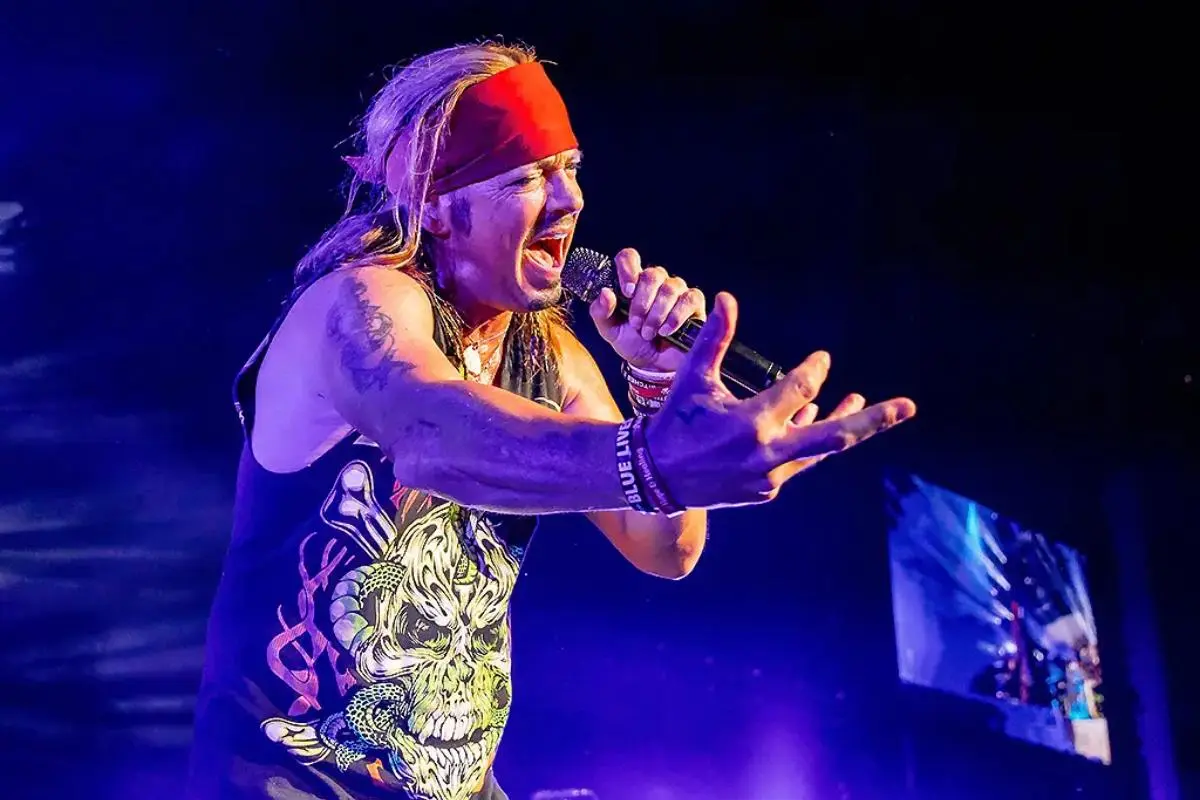 Jack Bruce wearing a red bandana sings passionately into a microphone, reaching toward the audience under blue stage lights.