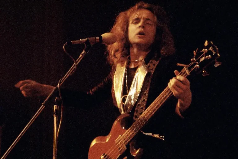 Jack Bruce sings into a microphone while playing bass guitar under warm stage lighting.
