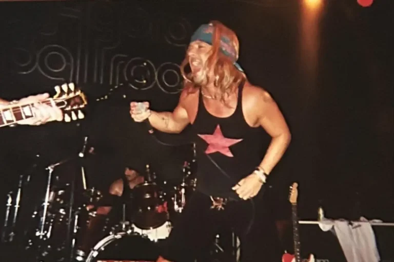 Jack Bruce performs energetically onstage in a sleeveless shirt, mid-gesture during a live concert.