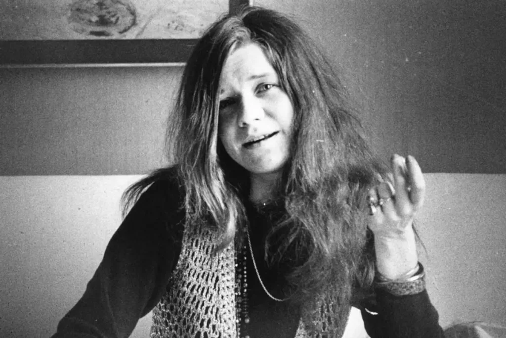 Janis Joplin with long, tousled hair smiles softly at the camera, raising one hand in a casual, relaxed gesture.