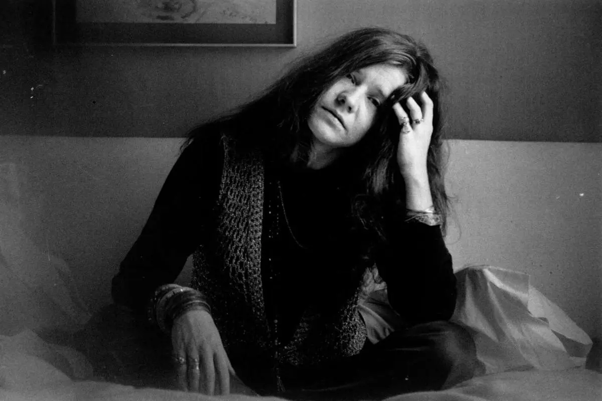 Janis Joplin sitting on a bed in a dimly lit room.