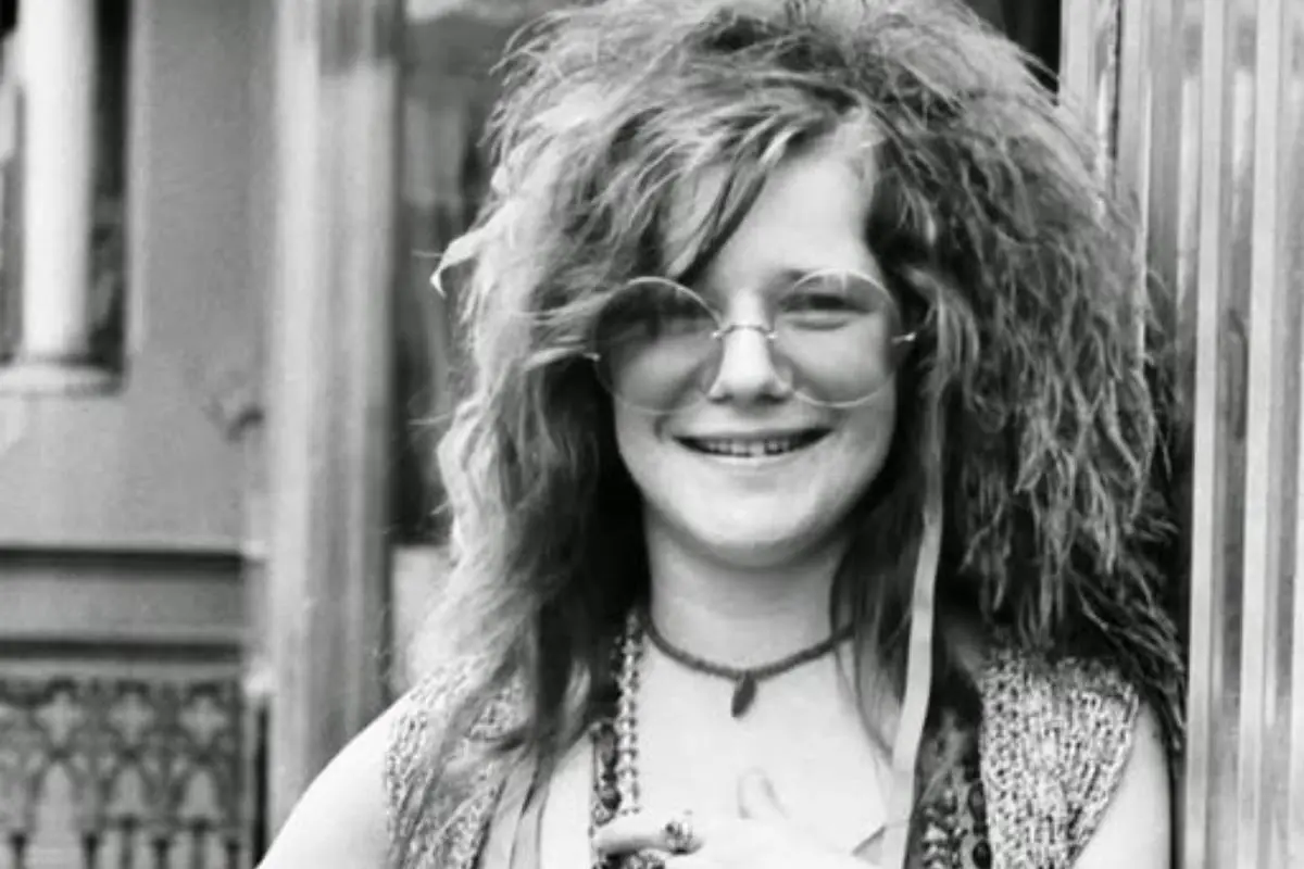 Janis Joplin smiling warmly while standing outdoors, wearing round glasses and a beaded necklace.