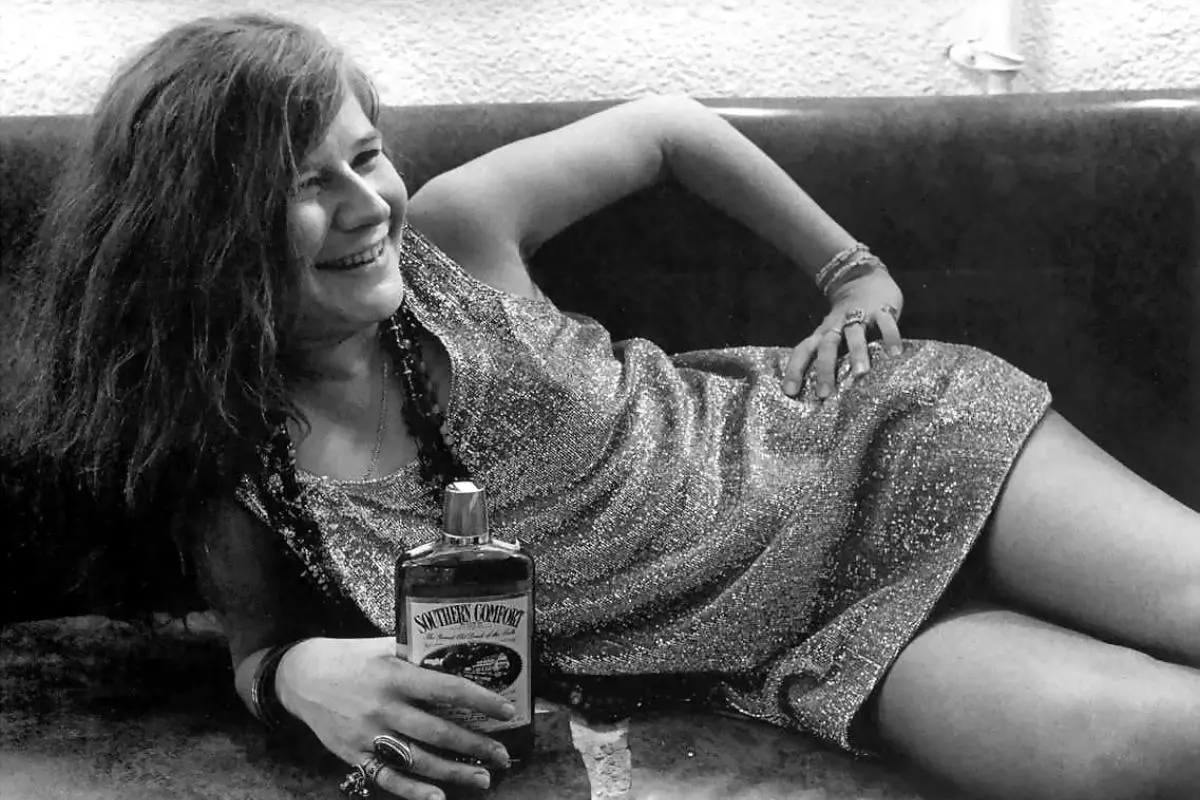 Janis Joplin reclining on a couch, laughing while holding a bottle.
