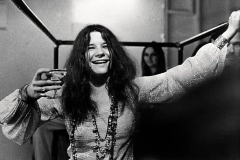 Janis Joplin backstage, smiling and raising a drink in one hand.