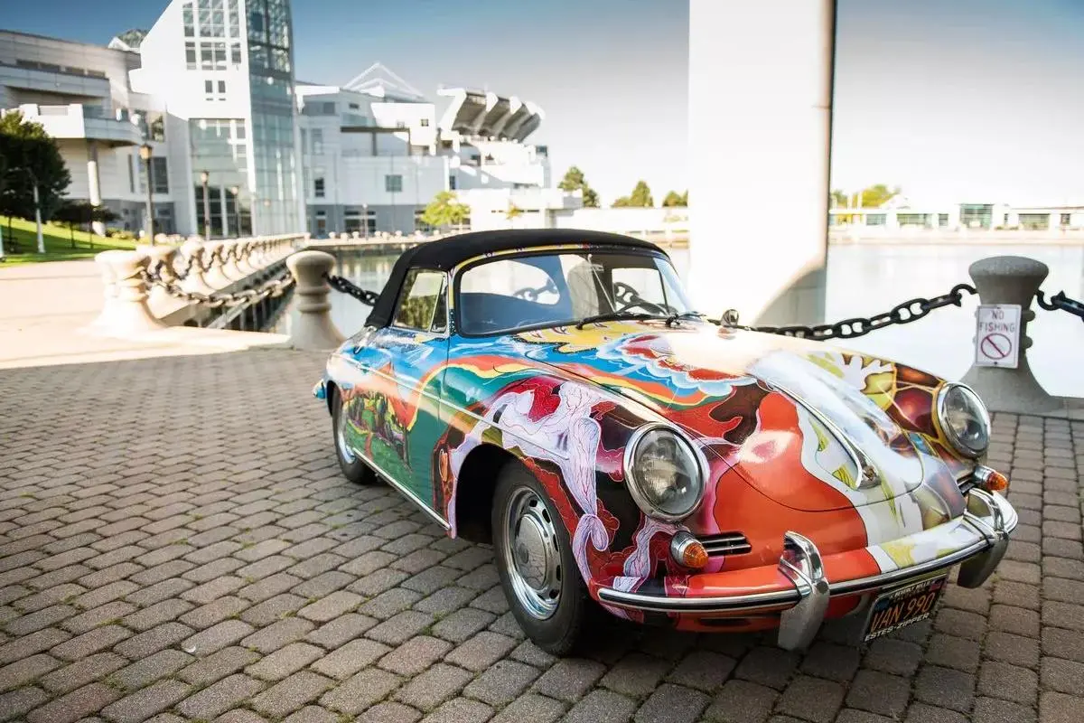 Janis Joplin’s Porsche sports car covered in vibrant psychedelic artwork is parked along a waterfront promenade near modern buildings.