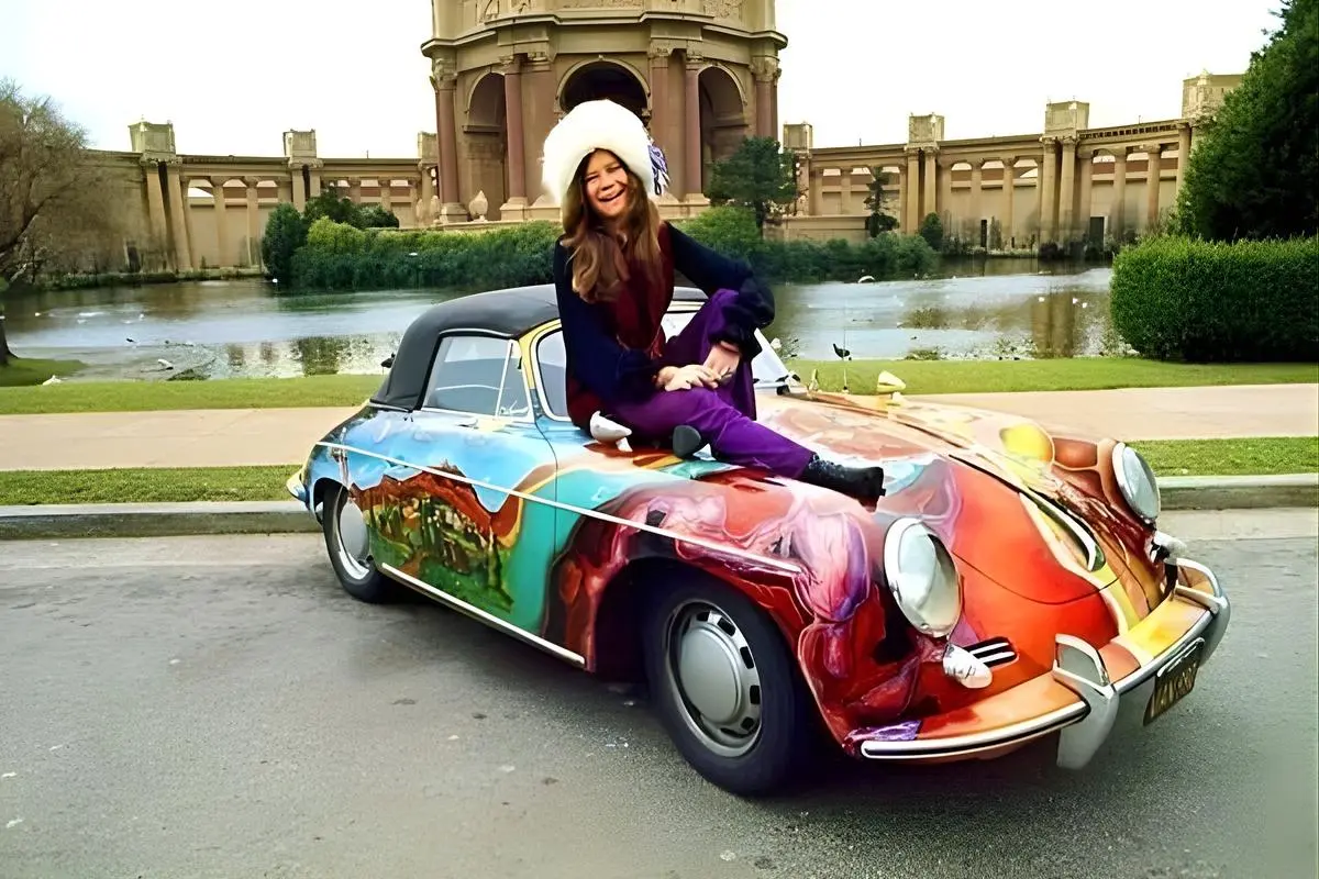 Janis Joplin in colorful 1960s-style clothing sits on the hood of a brightly painted psychedelic Porsche with a grand architectural building in the background.
