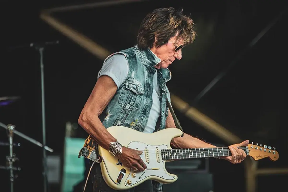 Jeff Beck playing electric guitar live on stage, wearing a sleeveless denim vest.