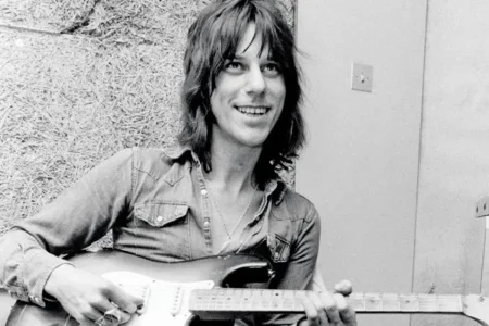 Jeff Beck on Hendrix & McLaughlin: The Guitar That Screamed For Him Jeff Beck sits casually with an electric guitar, smiling while holding the instrument.