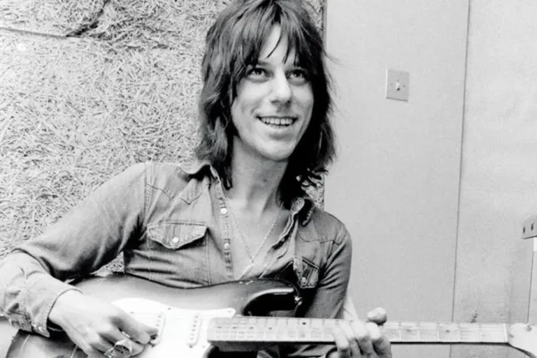 Jeff Beck sits casually with an electric guitar, smiling while holding the instrument.
