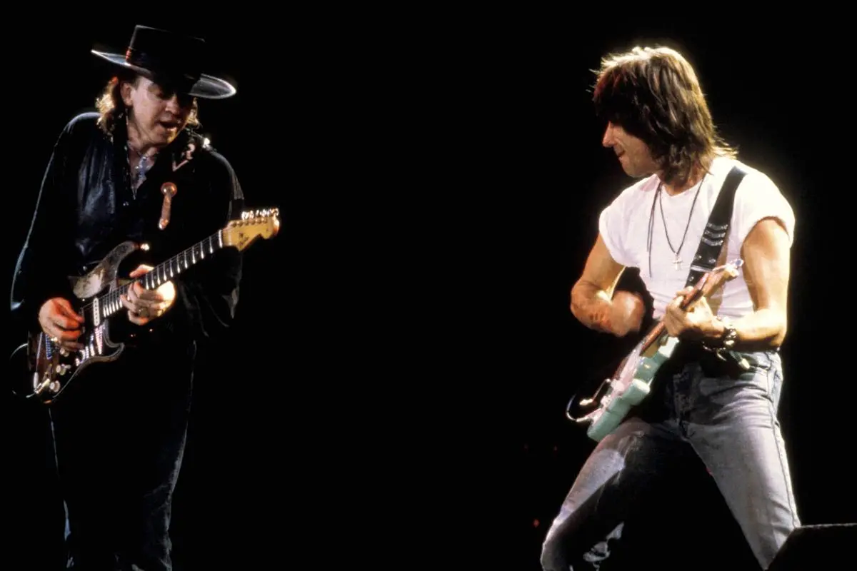 Stevie Ray Vaughan and Jeff Beck face each other onstage, each intensely playing electric guitar against a dark background.