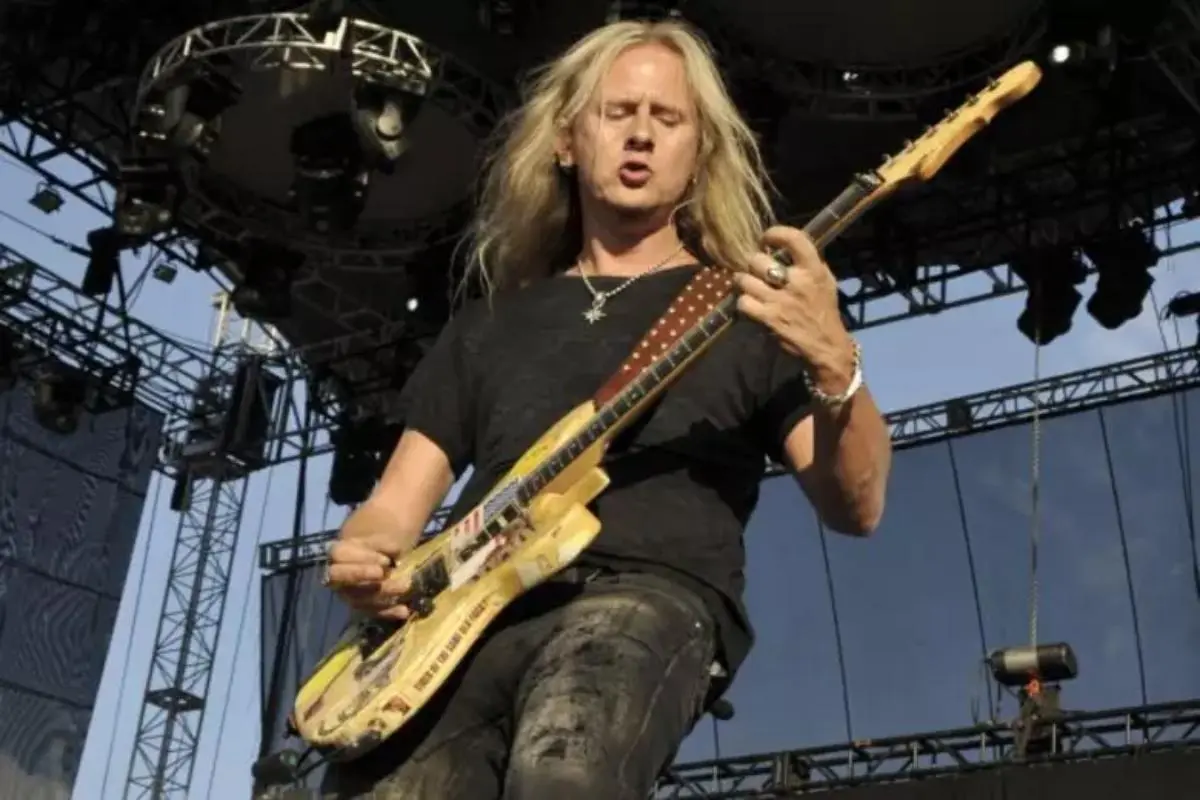 Jerry Cantrel performs on an outdoor stage, intensely playing an electric guitar decorated with stickers.