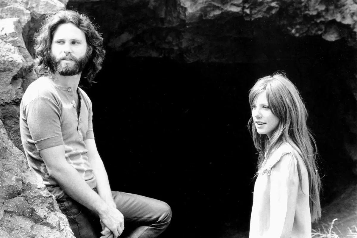 Jim Morrison and Pamela Courson standing together near a rocky cave entrance.