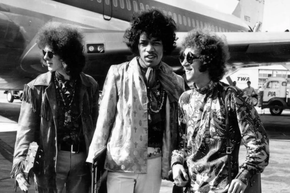 Jimi Hendrix with bandmates stand on an airport tarmac, wearing patterned 1960s clothing and smiling as they talk together.