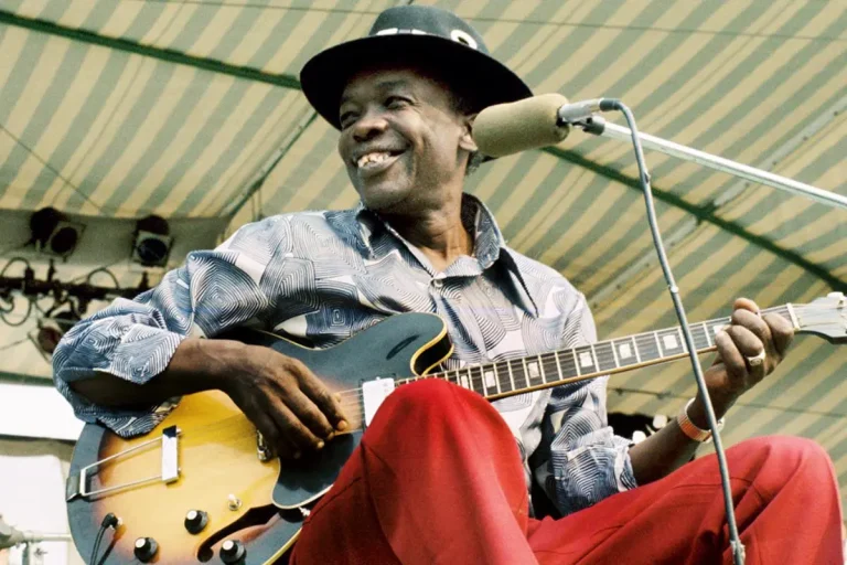 John Lee Hooker performing outdoors onstage, smiling while seated with an electric guitar.