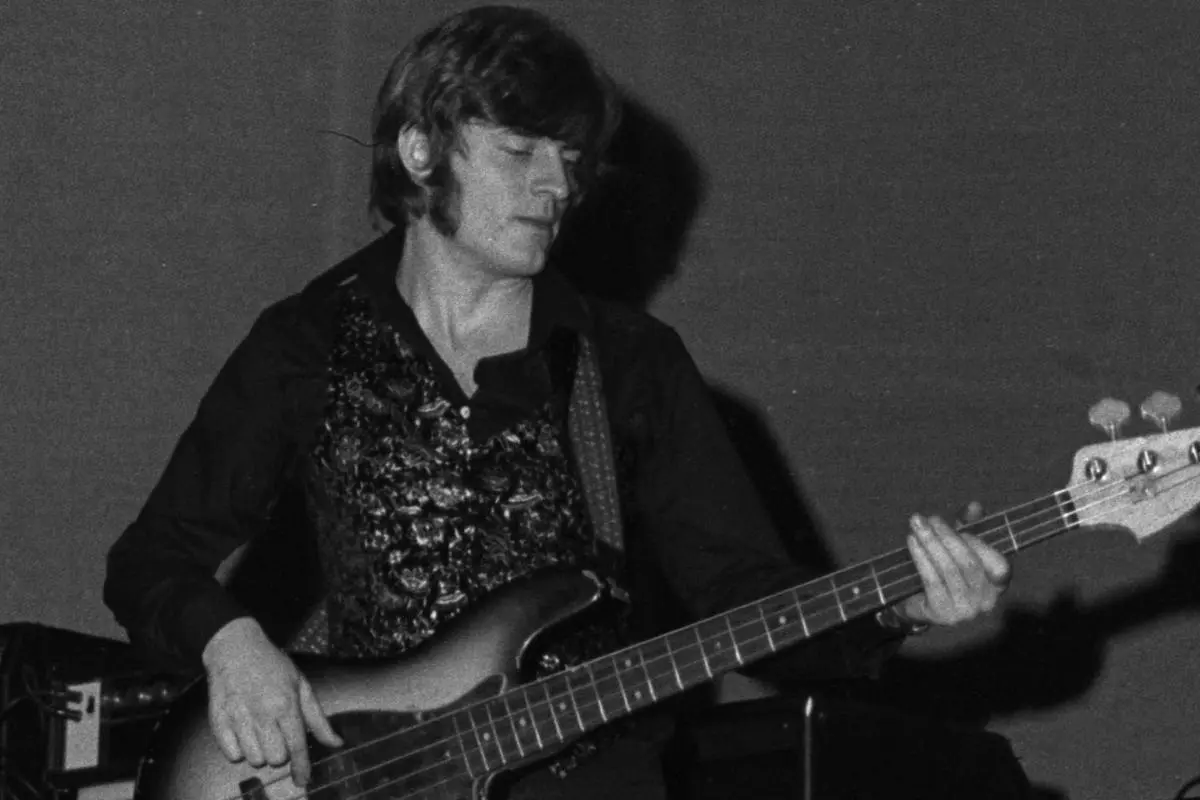 John Paul Jones plays a bass guitar with eyes closed, wearing a dark shirt and patterned vest under stage lighting.