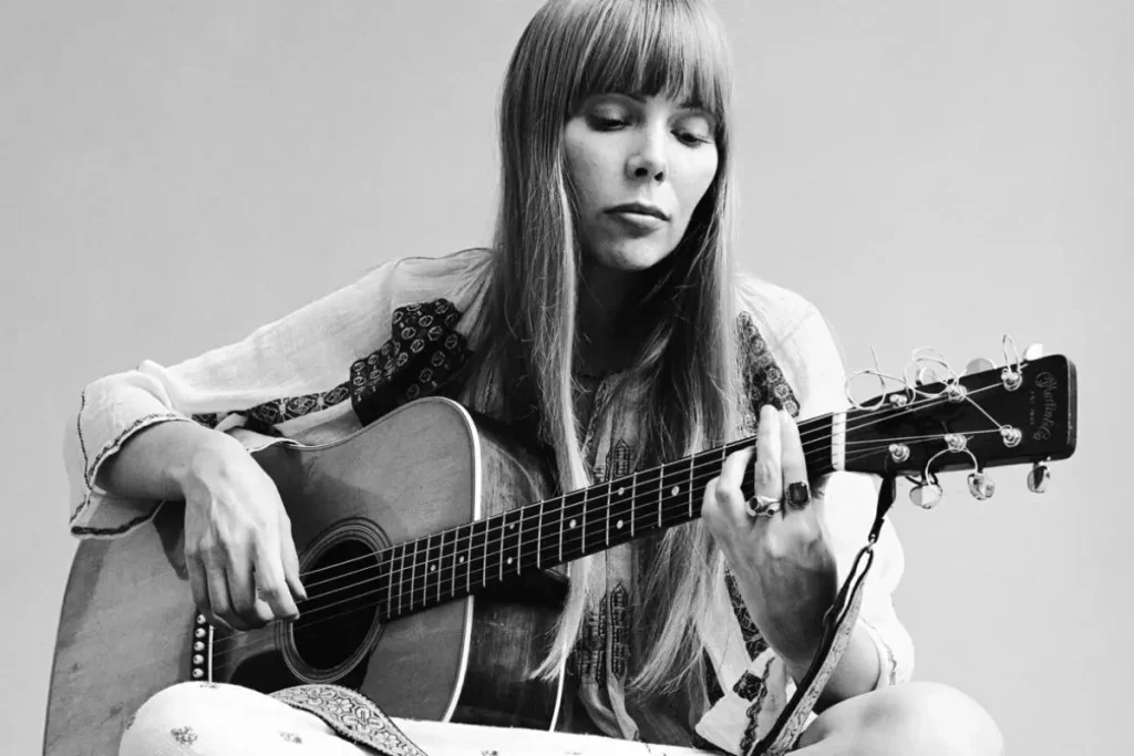 Joni Mitchell seated and playing an acoustic guitar.