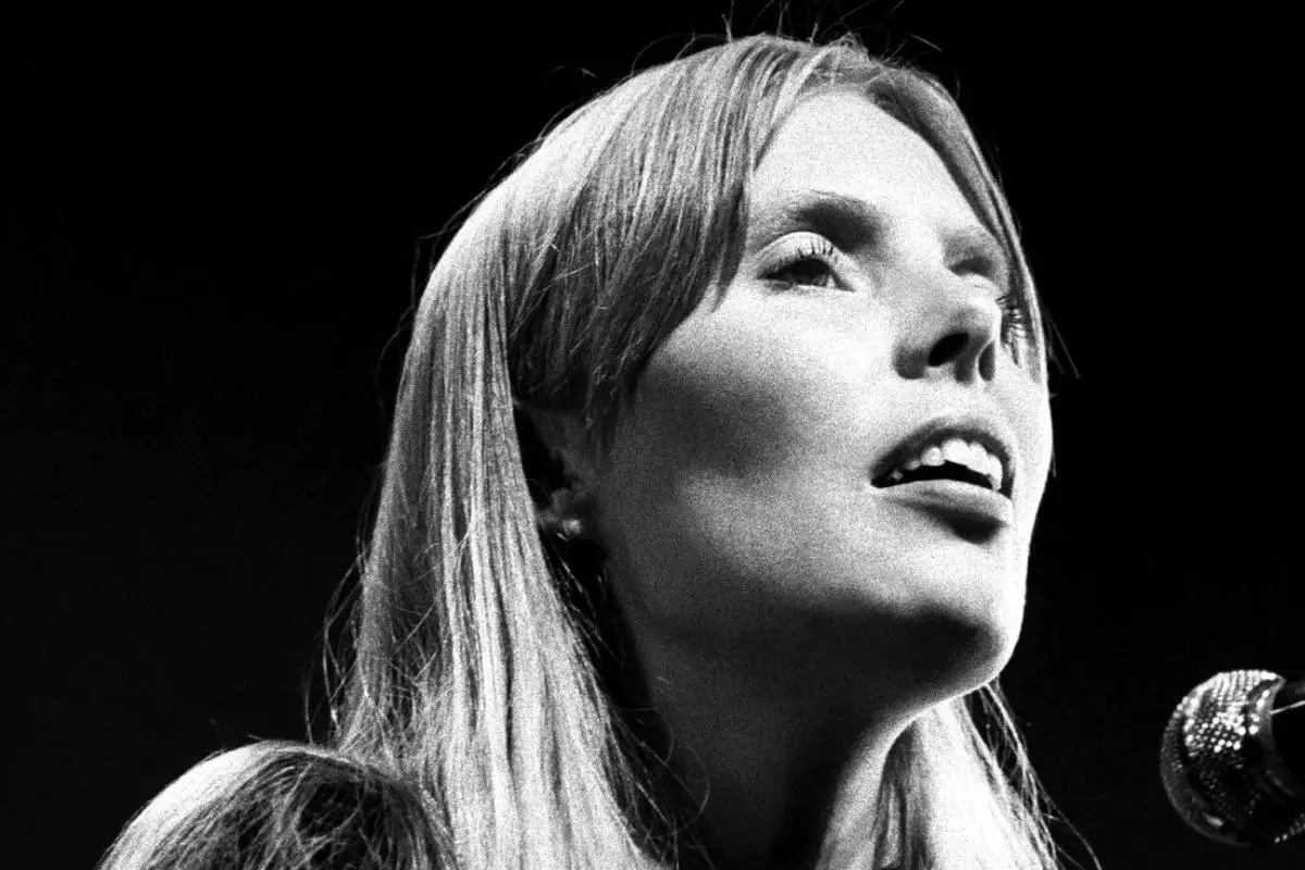 Joni Mitchell performs under a spotlight, looking upward as she sings into a microphone.