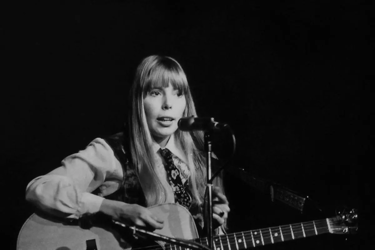 Joni Mitchell performs on stage with an acoustic guitar, singing into a microphone.