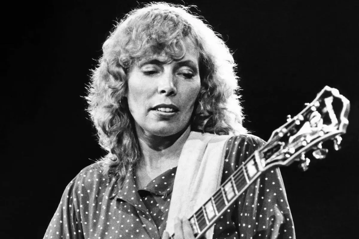 Joni Mitchell performing on stage, wearing a patterned shirt.