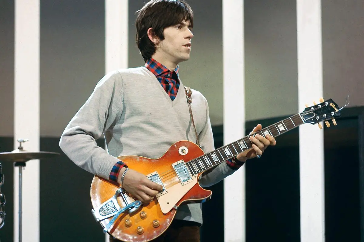 Keith Richards plays an electric guitar onstage, wearing a sweater over a collared shirt under bright studio lighting.