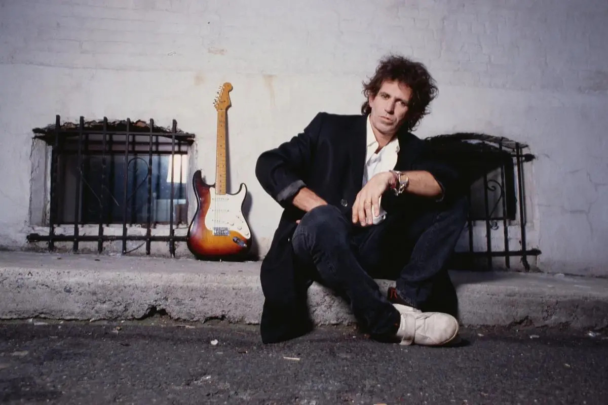 Keith Richards sits on a curb against a worn white wall, dressed in a black blazer and white shirt.