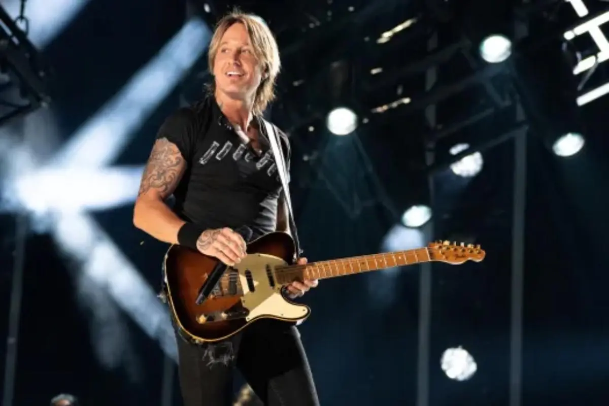 Keith Urban performs on a brightly lit stage, smiling while holding an electric guitar.