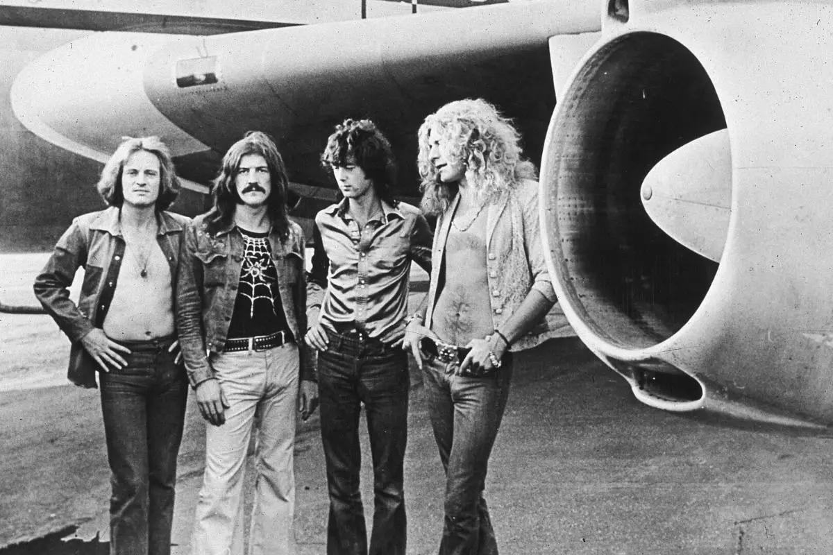 Four members of Led Zeppelin stand on an airport tarmac beside the engine of their private tour plane.