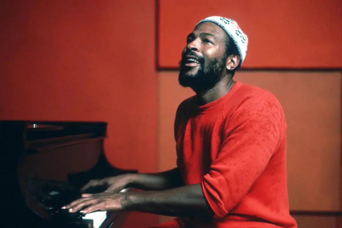 Marvin Gaye sits at a piano wearing a red long-sleeve shirt and a knitted beanie.