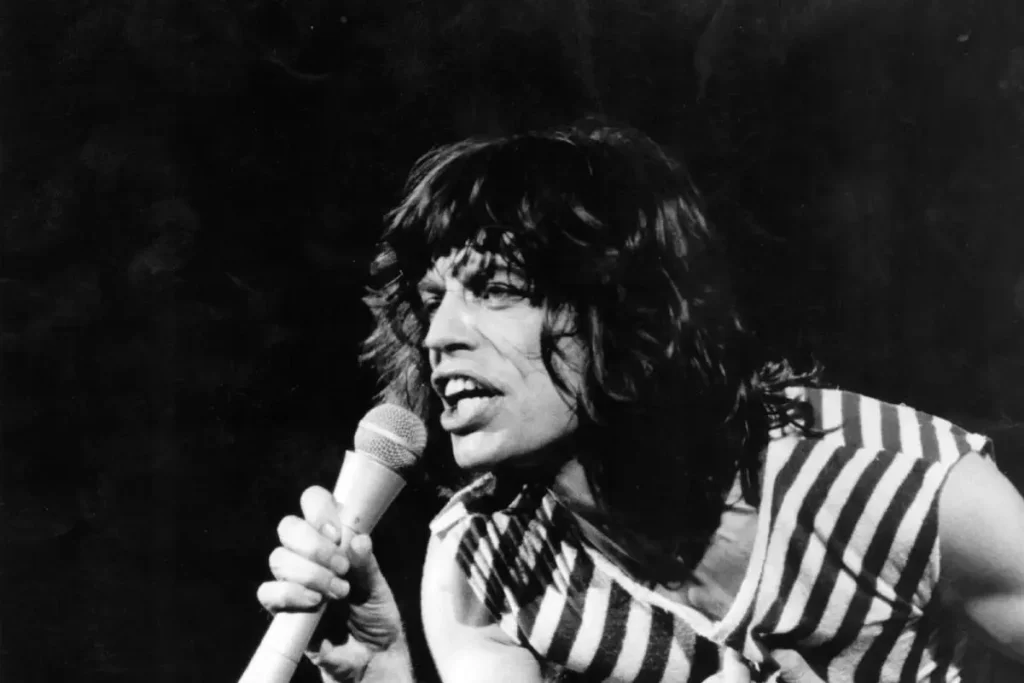 Black-and-white photo of Mick Jagger singing into a microphone during a live performance.