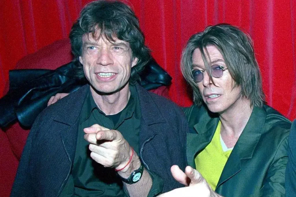 Mick Jagger & David Bowie in 1973: Glam’s Most Dangerous Friendship Mick Jagger and David Bowie sit closely together against a red backdrop, one pointing toward the camera while both appear mid-conversation.