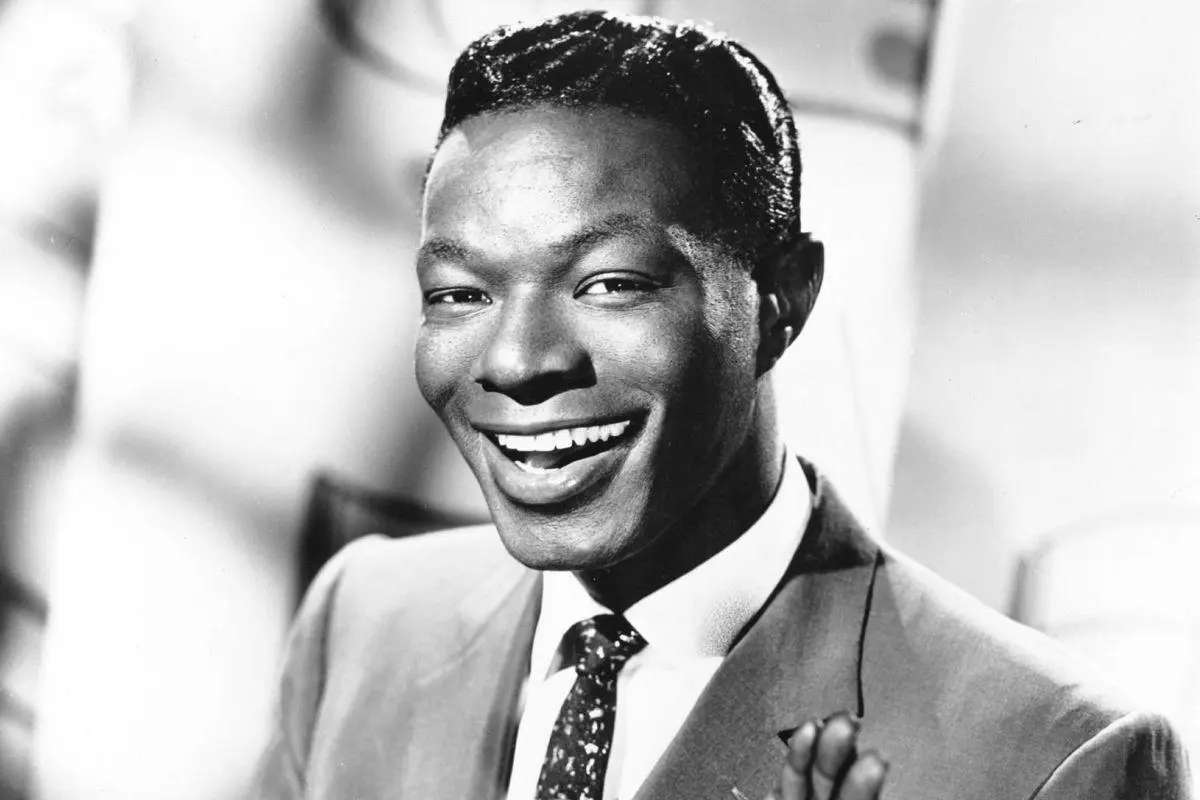 Nat King Cole smiling warmly, wearing a suit and tie with his hair neatly styled, looking slightly to the side.